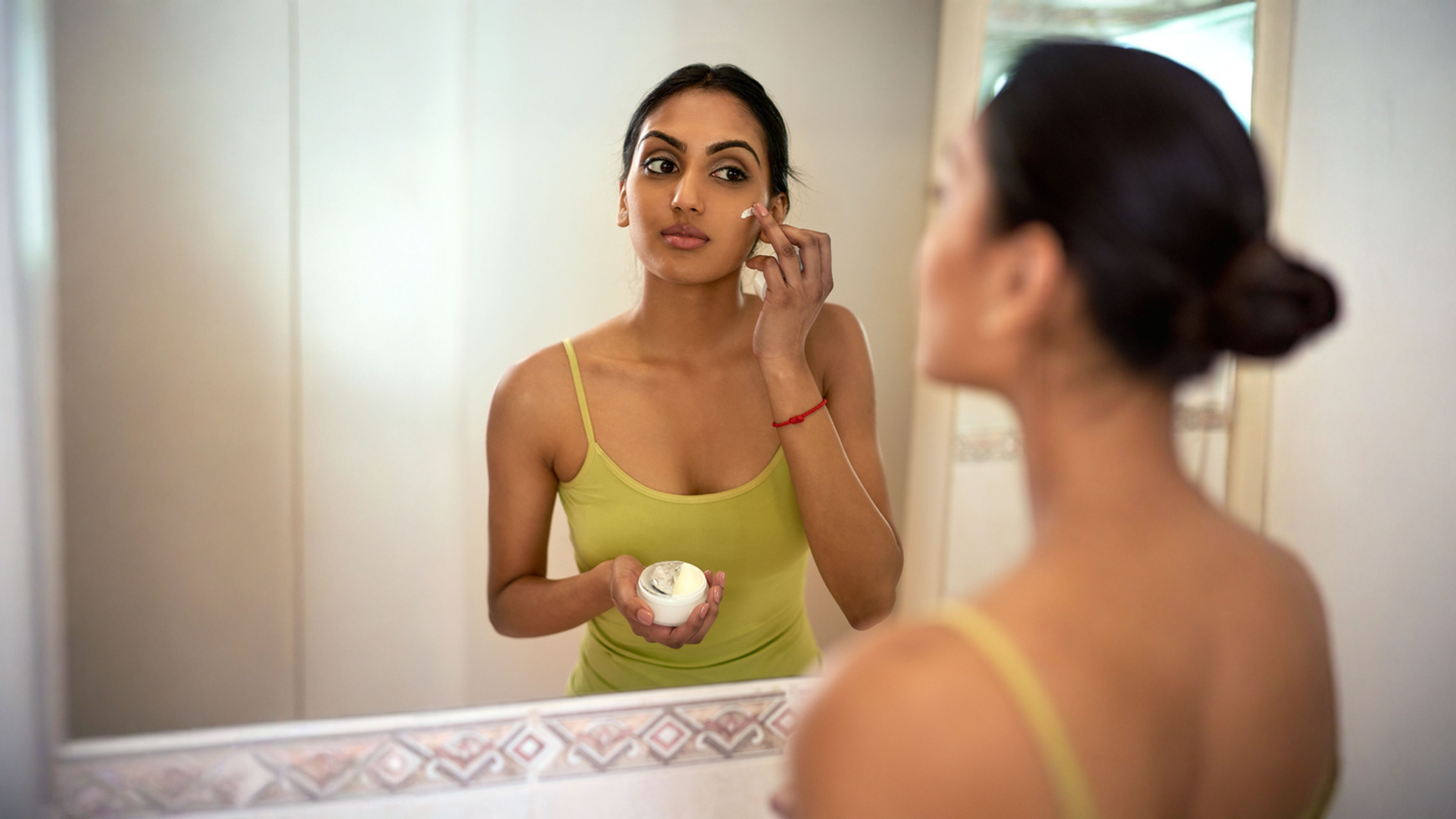Woman in green dress applying skincare essentials Infront of a mirror.