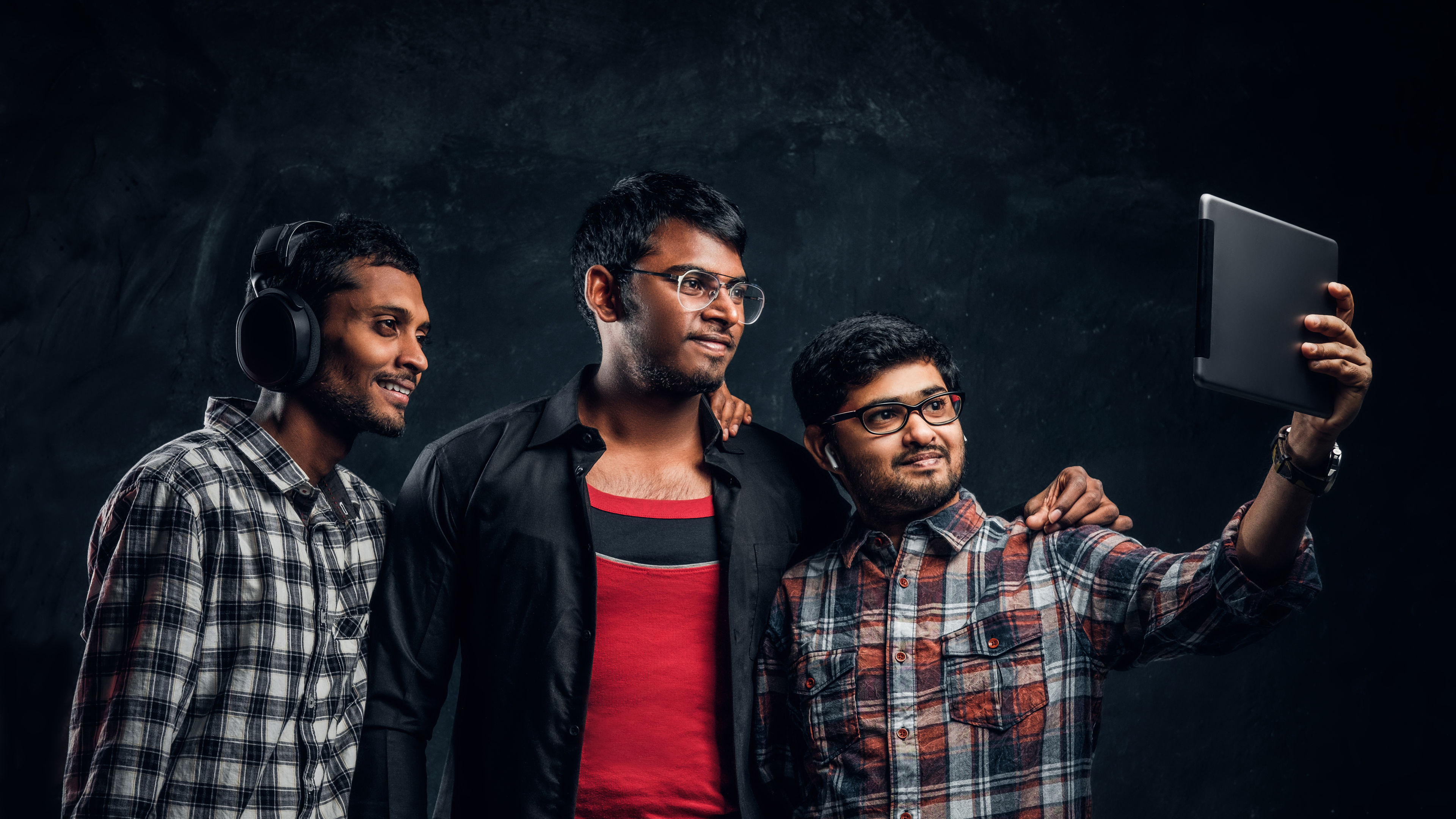 
Three happy Indian friends in casual clothes stand in a hug and taking selfie on his tablet